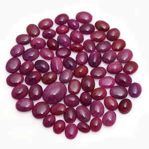 May include: A collection of oval-shaped, polished ruby gemstones in varying shades of red and pink. The gemstones are arranged closely together, showcasing their smooth surfaces and natural colour variations. The gemstones are a deep red colour.