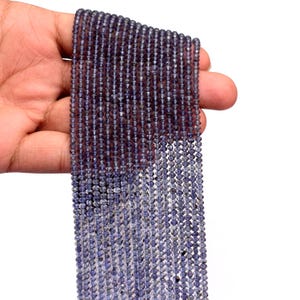 Pode incluir: Fio de pequenas contas facetadas azul escuro. As contas são enfiadas, criando um efeito de cascata. As contas são seguradas em uma mão contra um fundo branco. As contas têm um brilho sutil.