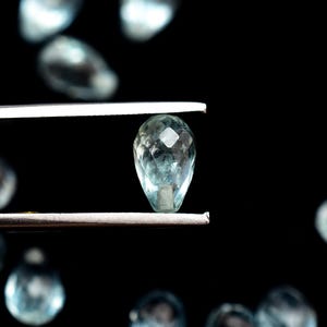 Può includere: Una gemma di acquamarina sfaccettata a forma di goccia è tenuta da una pinzetta su uno sfondo nero. La gemma è di colore azzurro pallido e ha molteplici sfaccettature che catturano la luce. Altre gemme sfocate sono sparse sullo sfondo.