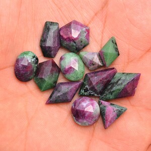 May include: A collection of faceted ruby gemstones in various shapes, including oval, hexagonal, and diamond. The gems display a mix of deep red and green hues, with visible internal patterns. The stones are displayed on a light-colored surface.