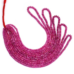 Può includere: Fili di piccole gemme rotonde, rosa intenso, probabilmente rubini, sono esposti su sfondo bianco. Le perle sono infilate su un filo rosso, creando un effetto a cascata. Le gemme hanno un aspetto lucido.