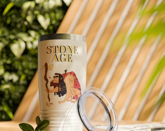 May include: A cream-colored tumbler with the words "STONE AGE" in gold lettering. The tumbler features a cartoon caveman illustration. A clear lid sits next to the tumbler. The tumbler is on a wooden surface.