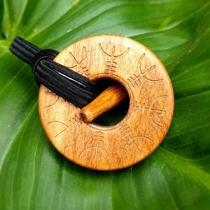 May include: A wooden hair stick with a carved circular design. The stick is threaded through a hole in the centre of the circle. The circle is decorated with a repeating pattern of carved lines.