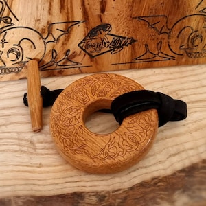 Könnte beinhalten: Ein Holz-Toggle-Verschluss mit einem Lebensbaum-Design. Der Verschluss ist aus hellbraunem Holz gefertigt und hat ein schwarzes Lederband. Der Verschluss befindet sich auf einer Holzoberfläche.