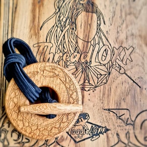 May include: A wooden hair tie with a carved design. The hair tie is in the shape of a circle with a smaller circle in the centre. The hair tie is brown and has a black elastic band attached to it.