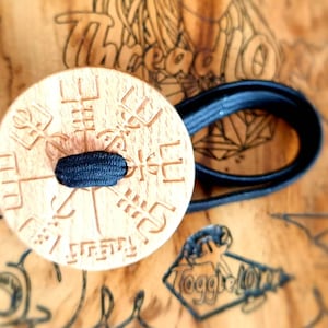 Puede incluir: Un botón de madera con una banda elástica negra. El botón tiene un diseño circular con un patrón tallado y el texto "Toggle Love" en la parte inferior.