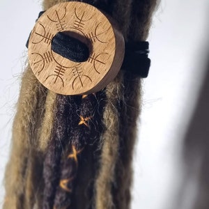 Op de afbeelding: Een houten haarband met een rond ontwerp en een zwart elastiek. De haarband is om bruin en blond dreadlocks gewikkeld.
