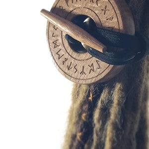 Könnte beinhalten: Ein Holzhaargummi mit einem runden Design, das einen geschnitzten Kreis mit Runen am Rand zeigt. Das Haargummi ist braun und hat ein schwarzes Gummiband.
