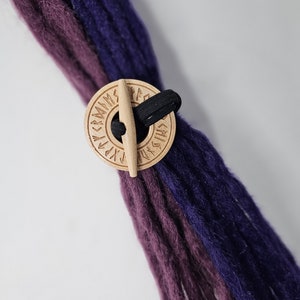 May include: A wooden round bead with runes carved into the surface. The bead is threaded onto a black elastic cord and is used to hold a bundle of purple and dark purple yarn.