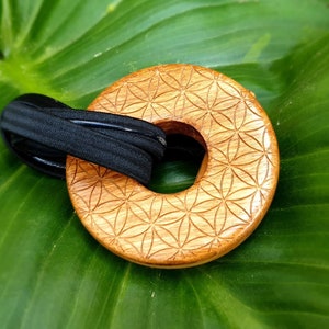 Op de afbeelding: Een houten haarband met een lasergesneden bloemenontwerp. De lichtbruine, cirkelvormige band heeft een zwarte elastische band. Het haaraccessoire ligt op een levendig groen blad.