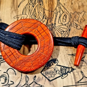 Puede incluir: Un botón de madera con un acabado rojo-marrón y un diseño geométrico. El botón está unido a un cordón negro y tiene un mango de madera.