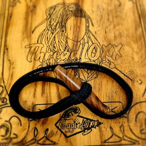 Może przedstawiać: Czarny elastyczny sznurek z drewnianym zapięciem toggle. Sznurek jest zapętlający, a zapięcie znajduje się w środku pętli. Tło to drewniana powierzchnia z wypalonymi w drewnie napisami "ThreadLove" i "ToggleLove".