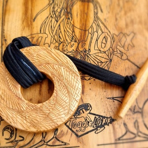 May include: A wooden pendant with a detailed, radial pattern, attached to a black cord with a wooden toggle. The pendant is on a wooden surface with the text "ThreadLoX" and "ToggleLoX" printed on it.