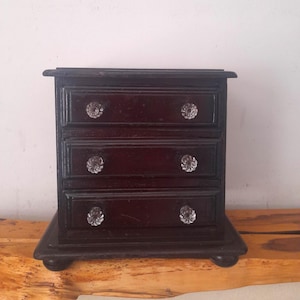 May include: A small, dark brown wooden chest of drawers featuring three horizontal drawers. Each drawer is equipped with two clear, faceted glass knobs. The unit stands on four rounded feet and has a smooth, polished finish on the front surface.