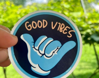 May include: A round sticker with a navy blue background and a light blue hand gesture. The words "GOOD VIBES" are in a peach color. The sticker has a white border and is held up against a blurred green background.