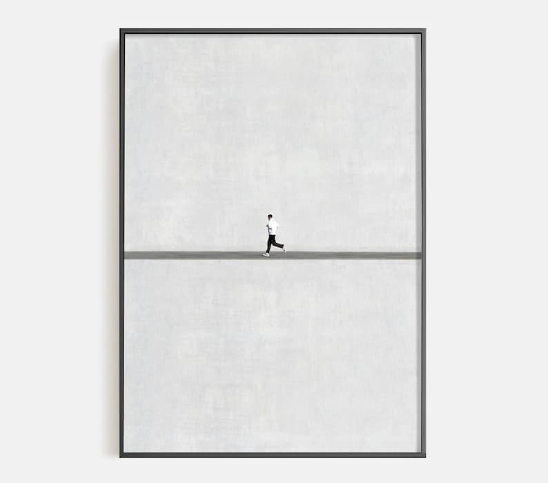 May include: Framed art print featuring a minimalist design. A small figure in black and white running across a gray line against a textured, light gray background. The artwork is encased in a black frame.