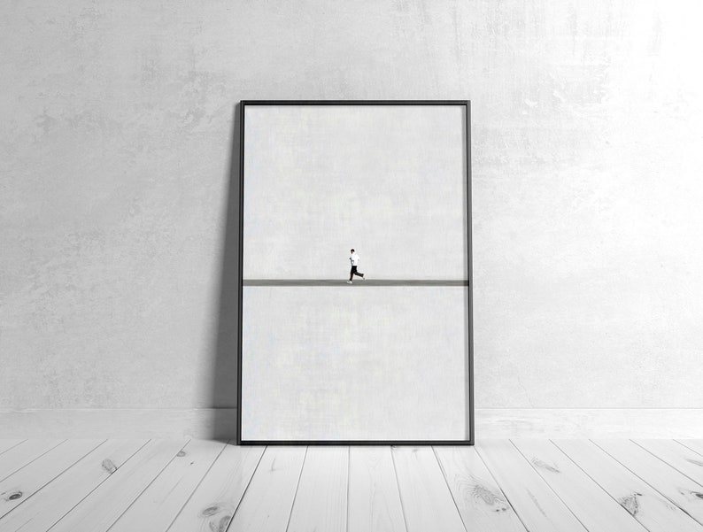 May include: Framed art print featuring a minimalist design. A small figure in black and white is running on a gray line against a white background. The artwork is encased in a black frame and leans against a white wall.