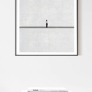 May include: A square framed wall art print featuring a minimalist composition of a small figure standing in the center of a horizontal line, dividing a textured light gray background into two equal sections, enclosed in a thin black frame.