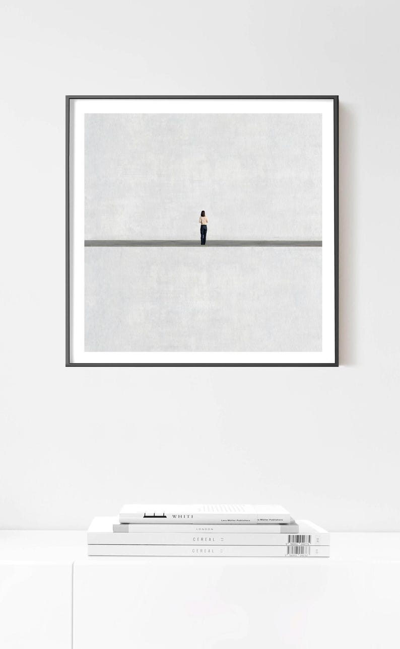 May include: A square framed wall art print featuring a minimalist composition of a small figure standing in the center of a horizontal line, dividing a textured light gray background into two equal sections, enclosed in a thin black frame.