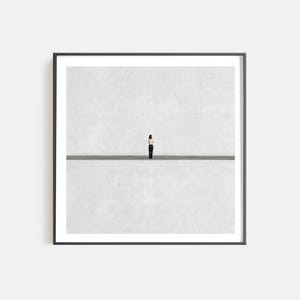 May include: A framed square wall art print featuring a small figure standing in the center of a vast, light gray space divided by a single, thin, dark horizontal line. The figure is wearing a light-colored top and dark trousers, facing away.