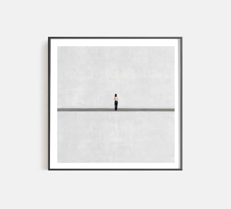 May include: A framed square wall art print featuring a small figure standing in the center of a vast, light gray space divided by a single, thin, dark horizontal line. The figure is wearing a light-colored top and dark trousers, facing away.
