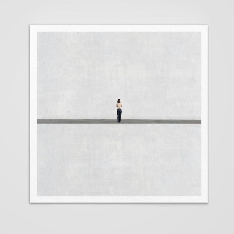 May include: A square art print featuring a small figure standing in the center of a vast, empty space. The composition is divided horizontally by a dark line, separating a textured light gray upper section from a matching lower section.
