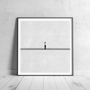 May include: A square framed wall art print featuring a minimalist composition with a small figure standing in the center of a horizontal line, set against a vast, light gray background that creates a sense of expansive, open space.