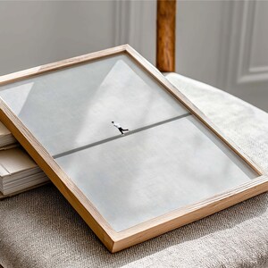 May include: A framed art print featuring a minimalist illustration of a figure walking a tightrope. The artwork is encased in a light-colored wooden frame with a clear glass cover. The frame rests on a stack of books and a neutral-toned upholstered surface.