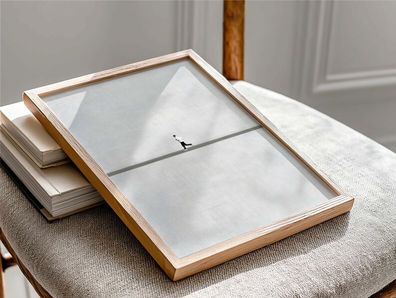 May include: A framed art print featuring a minimalist illustration of a figure walking a tightrope. The artwork is encased in a light-colored wooden frame with a clear glass cover. The frame rests on a stack of books and a neutral-toned upholstered surface.