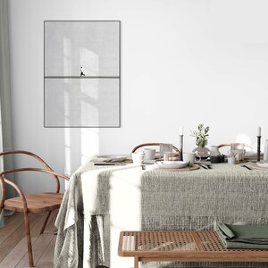 May include: A framed minimalist art print featuring a person walking on a line against a textured gray background. The artwork is displayed above a dining table set with tableware, candles, and a green folded cloth. A wooden chair and bench are in the foreground.