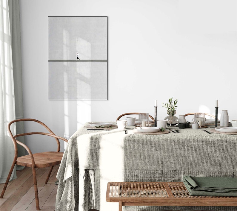 May include: A framed minimalist art print featuring a person walking on a line against a textured gray background. The artwork is displayed above a dining table set with tableware, candles, and a green folded cloth. A wooden chair and bench are in the foreground.