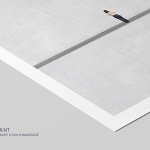 May include: A fine art print featuring a minimalist composition of a light gray surface divided by a dark gray line, with a small figure in a tan shirt and dark pants lying horizontally across the line. The text FINE ART PRINT ORIGINALFOTOGRAFIE EKA SHARASHIDZE is visible in the bottom left corner.