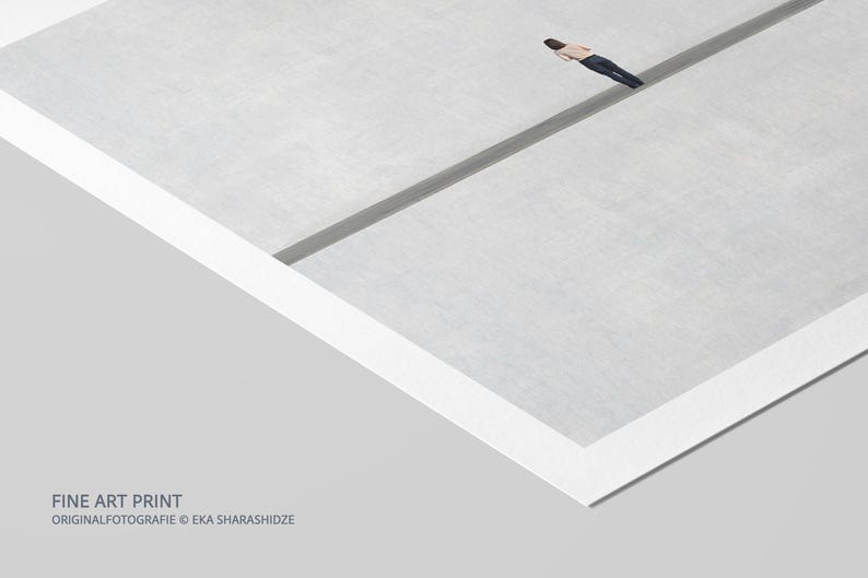 May include: A fine art print featuring a minimalist composition of a light gray surface divided by a dark gray line, with a small figure in a tan shirt and dark pants lying horizontally across the line. The text FINE ART PRINT ORIGINALFOTOGRAFIE EKA SHARASHIDZE is visible in the bottom left corner.