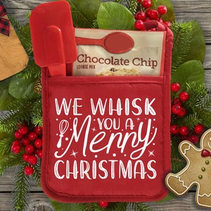 May include: Red oven glove with the text "We Whisk You a Merry Christmas" in white. The oven glove has a red spatula and a bag of chocolate chip cookie mix inside. A gingerbread man biscuit is on the right side of the image.