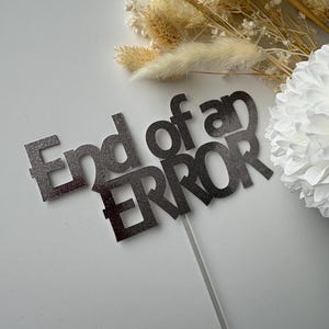 May include: A silver glitter cake topper with the words "End of an ERROR" on a clear stick. The topper is set against a white background with dried flowers and a white paper flower.
