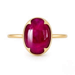 May include: A gold ring featuring a large, oval-shaped, deep magenta-colored cabochon gemstone set in a smooth bezel with four distinct prongs at the cardinal points. The band is thin, polished, and circular, providing a clean, simple frame.
