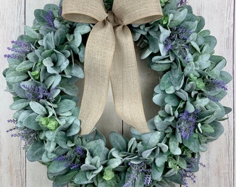 May include: A green and grey wreath with purple flowers and a brown burlap bow. The wreath is made of artificial greenery and flowers.
