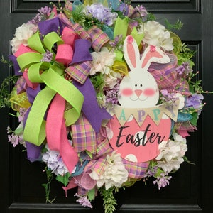May include: A pink and green Easter wreath featuring a bunny holding a sign that reads "Happy Easter". The wreath is decorated with flowers, ribbons, and mesh.