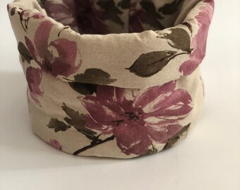 May include: A fabric storage basket with a floral design. The basket is beige with large, watercolour-style purple flowers and brown leaves. The top edge is folded over, revealing the floral pattern on the inside.
