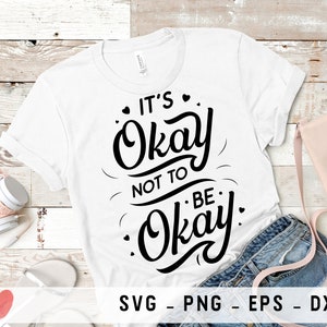 以下が含まれることがあります： 白いTシャツに黒い文字で「It's Okay Not To Be Okay」と書かれています。Tシャツは白い木製の表面に置かれ、ヘッドホンとピンクのハンドバッグが置かれています。