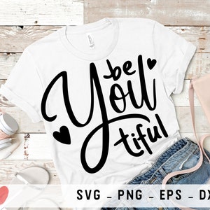 Könnte beinhalten: Weißes T-Shirt mit schwarzem Text, der "be you tiful" lautet, mit einem Herz-Motiv auf jeder Seite des Textes. Das Shirt liegt auf einer Holzoberfläche mit einer Jeans und einer Handtasche.