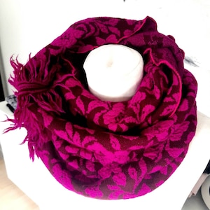 Könnte beinhalten: Ein magenta- und bordeauxfarbener Loop-Schal mit floralem Muster. Der Schal hat einen Fransenrand und ist über einer weißen Form drapiert. Das Design des Schals zeigt ein sich wiederholendes Muster aus Blättern.