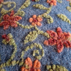 Könnte beinhalten: Nahaufnahme eines blauen Textils mit einem gestickten Blumenmuster. Die Stickerei zeigt orange und rote Blüten mit grünen Blättern und Stielen. Die Textur des Stoffes und der Stickerei ist sichtbar.