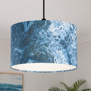 Może przedstawiać: Cylindryczna lampa wisząca z abażurem w kształcie bębna, ozdobionym niebiesko-białym abstrakcyjnym wzorem przypominającym wirującą wodę morską lub pianę. Abażur zawieszony jest na cienkim czarnym przewodzie przymocowanym do centralnego punktu.