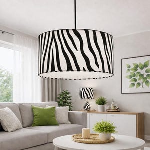 Może przedstawiać: Cylindryczna lampa wisząca typu bęben z abażurem z tkaniny w czarno-biały wzór w paski zebry. Lampa wisi na cienkim czarnym pręcie i posiada biały panel dyfuzyjny na spodzie, który zmiękcza emitowane światło.