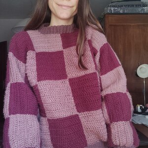 K&ouml;nnte beinhalten: Ein pinkfarbener und burgunderfarbener kariert gestrickter Pullover mit langen &Auml;rmeln.