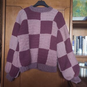 K&ouml;nnte beinhalten: Ein geh&auml;kelter Pullover mit einem karierten Muster in Lila- und Rosat&ouml;nen.
