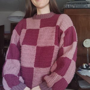 K&ouml;nnte beinhalten: Ein pinkfarbener und burgunderfarbener kariertes Strickpullover mit langen &Auml;rmeln und einem runden Ausschnitt.