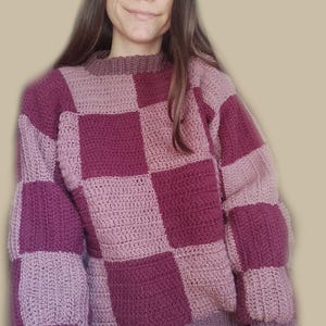 K&ouml;nnte beinhalten: Ein geh&auml;kelter Pullover mit einem karierten Muster in Rosa- und Lilat&ouml;nen.