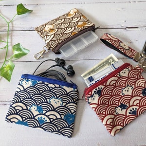 May include: Three small zippered pouches with a cat and wave pattern. The pouches are in blue, red, and brown. The pouches are on a white wooden surface.